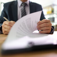 Businessman flips page of contract signing with pen after reading. Man in classic suit puts signature on paper sheet making successful deal